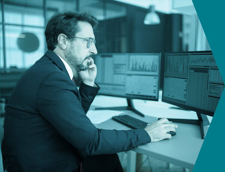 Man sitting at his desk with two computer screens, poring over the data, graphs and charts in front of him.